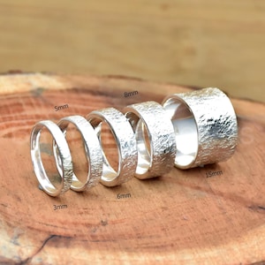 Könnte beinhalten: Fünf silberne Ringe in verschiedenen Breiten, von 3 mm bis 15 mm. Jeder Ring hat eine strukturierte Oberfläche. Die Ringe werden auf einer Holzoberfläche präsentiert. Die Breiten sind in Millimetern angegeben.
