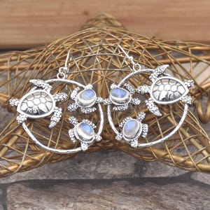 May include: Silver earrings with a sea turtle design. Each earring has a large turtle at the top and three smaller turtles surrounding a moonstone. The earrings are displayed on a gold star-shaped decorative object.