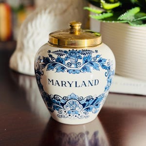 May include: A white ceramic jar with a gold lid, featuring blue floral patterns and the word "MARYLAND" in blue lettering. The jar is on a dark surface, with a blurred background.