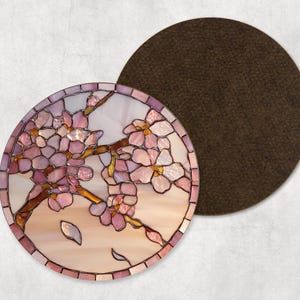 May include: A circular stained glass coaster with a cherry blossom design. The design features pink and white flowers on brown branches. A separate brown coaster is next to the glass coaster. The coaster is a decorative item.