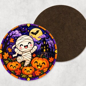 May include: A round coaster featuring a stained-glass style illustration of a happy mummy character surrounded by three smiling jack-o'-lanterns, autumn leaves, a haunted house, and bats against a full moon, with a dark brown circular backing.