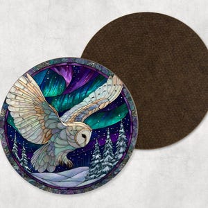 May include: Circular coaster depicting a white owl in flight, set against the Northern Lights. The design includes a snowy landscape with evergreen trees and a starry night sky. A separate brown coaster is also visible.