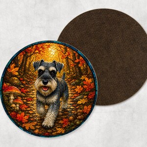 May include: A circular drinks coaster featuring a detailed illustration of a Schnauzer dog walking through an autumn forest with orange and red maple leaves, mushrooms, and trees. The coaster has a dark brown, textured felt backing for stability.