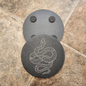 May include: Two round, dark grey slate coasters. One coaster features a detailed etched design of a snake with crescent moons and stars. The other coaster has two small, round, dark grey magnets attached. Coasters protect surfaces from heat and moisture.