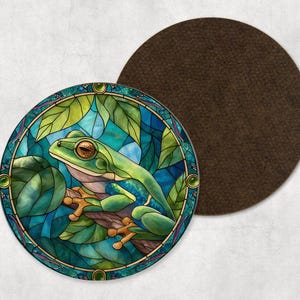 May include: A circular coaster with a stained-glass effect depicting a green frog on a branch with leaves. The design has a teal and blue background. The coaster has a brown backing.