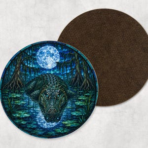 May include: A round drink coaster featuring a dark swamp scene with a green alligator resting in water under a full moon. The artwork has a stained glass aesthetic with deep blue and green tones. The reverse side is a solid dark brown felt material.