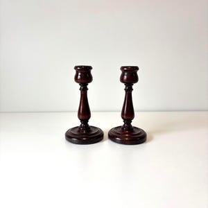 May include: A pair of dark brown wooden candlesticks. Each candlestick features a round base, a slender stem, and a flared top. The candlesticks are presented on a white surface.