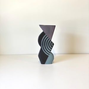 May include: A contemporary ceramic vase with a geometric design. The vase has a triangular top, a curved middle section with concentric circles, and a light blue base. The colour scheme includes shades of grey, black, and blue.