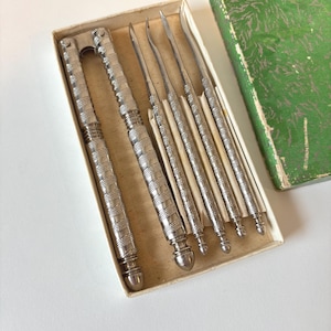Peut inclure: Un ensemble de casse-noix vintage. Le casse-noix et les pics argentés sont dans une boîte en carton avec un couvercle vert à motifs. Les outils ont des poignées texturées. L'ensemble est conçu pour casser les noix.