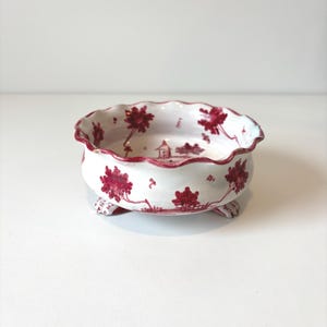 May include: A small, white ceramic bowl with a scalloped edge and three small feet. The bowl features a repeating pattern of red floral and landscape designs. The interior of the bowl is white.