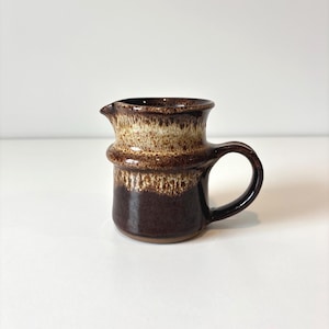 May include: A small, brown ceramic jug with a handle and spout. The jug has a dark brown base that fades to a lighter brown and tan speckled band around the middle. The rim is dark brown.