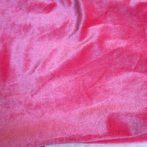 Puede incluir: Primer plano de una vibrante tela de terciopelo rosa. El material tiene una textura suave y afelpada con pliegues visibles y sutiles variaciones de color, creando una apariencia rica y lujosa. El brillo de la tela capta la luz, resaltando su profundidad.