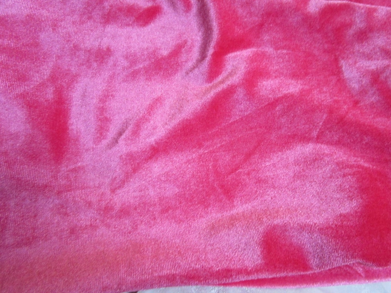 Puede incluir: Primer plano de una vibrante tela de terciopelo rosa. El material tiene una textura suave y afelpada con pliegues visibles y sutiles variaciones de color, creando una apariencia rica y lujosa. El brillo de la tela capta la luz, resaltando su profundidad.