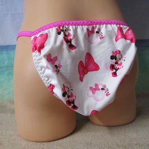 May include: White underwear with a pink trim and a pattern featuring Minnie Mouse and pink bows. The underwear is made of a soft fabric and has a string-style design.