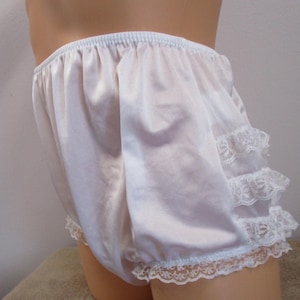 May include: White satin knickers with an elasticated waistband and ruffled lace trim. The vintage undergarment features a soft, flowing fabric and delicate lace detailing along the leg openings.