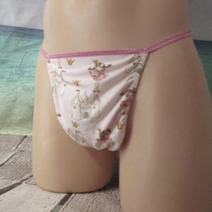May include: A light pink thong with a pink trim. The thong features a repeating pattern of castles, crowns, and princesses. The fabric is soft and lightweight, suitable for everyday wear.