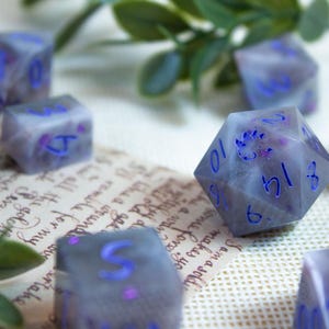 May include: A collection of polyhedral dice in various shapes, including D20s and D6s. The dice are a translucent gray with blue numbering and accents. Some dice have a floral design. The dice are scattered on a surface with handwritten text.