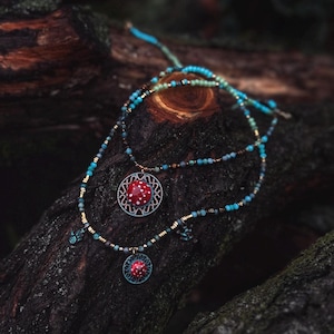 May include: Two layered necklaces with turquoise and gold beads. The necklaces feature pendants with red, dotted designs. The pendants are set in ornate, teal-colored metalwork. The necklaces are displayed on a dark, textured wooden surface.