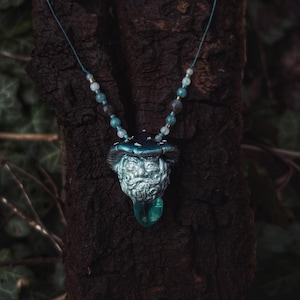 May include: A silver and blue necklace with a mushroom pendant featuring a face and a green gem. The necklace is made of beads and is hanging from a tree branch.