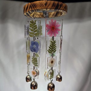 May include: A handcrafted wind chime featuring a wooden top and clear acrylic panels. Each panel showcases pressed flowers and ferns. Silver bells and a crystal prism hang below, creating a decorative and natural aesthetic.
