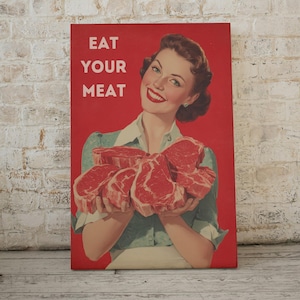 May include: A vintage-style canvas print with a red background and the text "EAT YOUR MEAT" in white. The image features a smiling woman holding several raw steaks. The woman is wearing a green shirt and white apron.