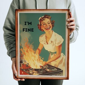 May include: A framed vintage-style poster with a woman in a white dress and apron standing in front of a stove with flames coming from a pan. The text on the poster reads "I'm Fine".