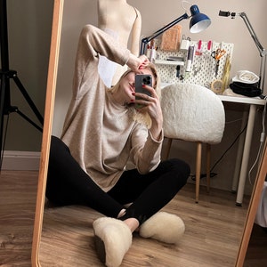 May include: A person wearing a beige jumper, black leggings, and white fluffy slippers is sitting on a wooden floor in front of a mirror. The person is taking a selfie with a smartphone.