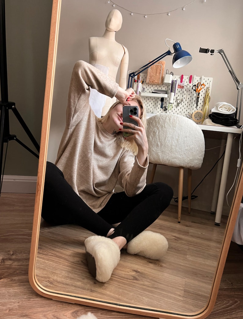 May include: A person wearing a beige jumper, black leggings, and white fluffy slippers is sitting on a wooden floor in front of a mirror. The person is taking a selfie with a smartphone.