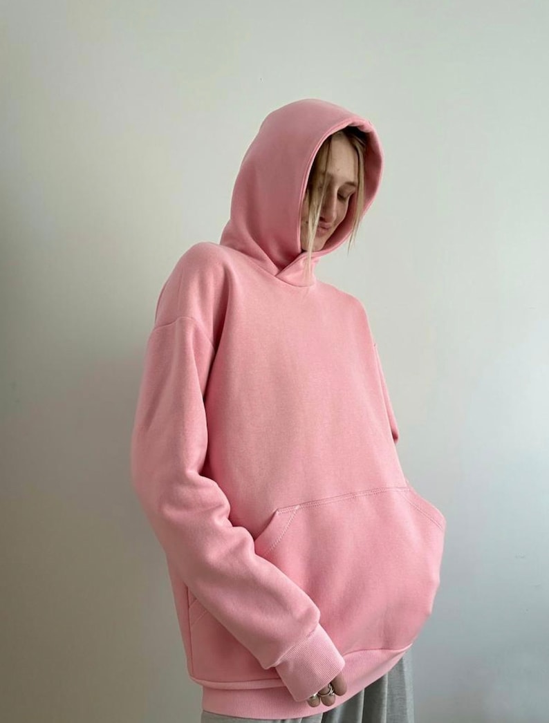 May include: A light pink hoodie with a large front pocket and a hood. The sweatshirt is made of a soft material and has ribbed cuffs and hem. The hoodie is a solid color and has a relaxed fit.