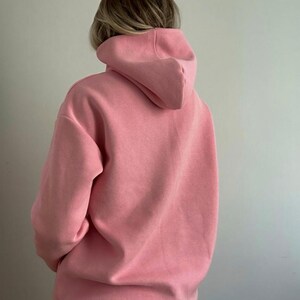 May include: A light pink hoodie with a large hood. The hoodie is made of a soft material and has a relaxed fit. The back of the hoodie is visible, showing the hood and the shoulders.