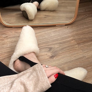 May include: A pair of white sheepskin slippers with black elastic straps. The slippers are being worn by a person with red nail polish and a ring on their finger.