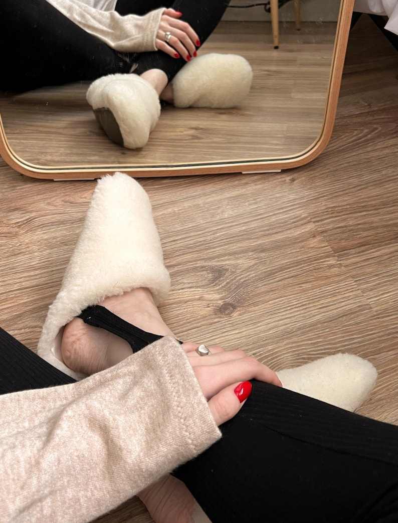 May include: A pair of white sheepskin slippers with black elastic straps. The slippers are being worn by a person with red nail polish and a ring on their finger.