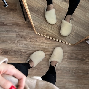 May include: A pair of white fluffy slippers with a black and white patterned design. The slippers are being worn by a person wearing black leggings and a beige sweater.