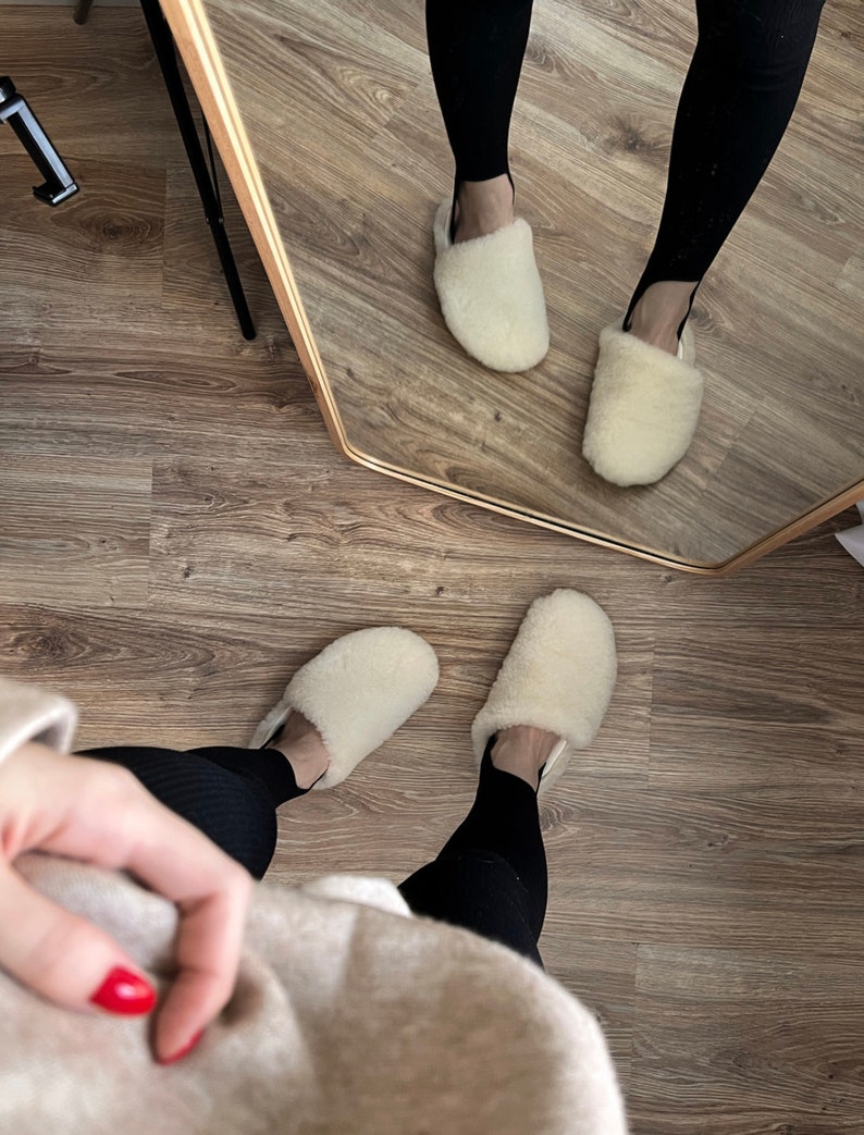 May include: A pair of white fluffy slippers with a black and white patterned design. The slippers are being worn by a person wearing black leggings and a beige sweater.