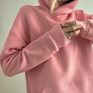 May include: A light pink hoodie with a kangaroo pocket and ribbed cuffs. The hoodie features a drawstring hood and a relaxed fit. The fabric appears to be a soft, comfortable material, perfect for casual wear.