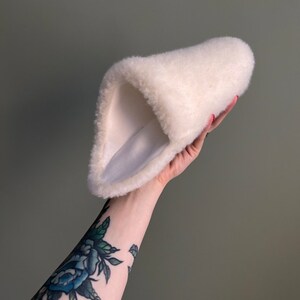 May include: A white fluffy slipper with a soft, plush lining. The slipper is folded in half and held in a hand.