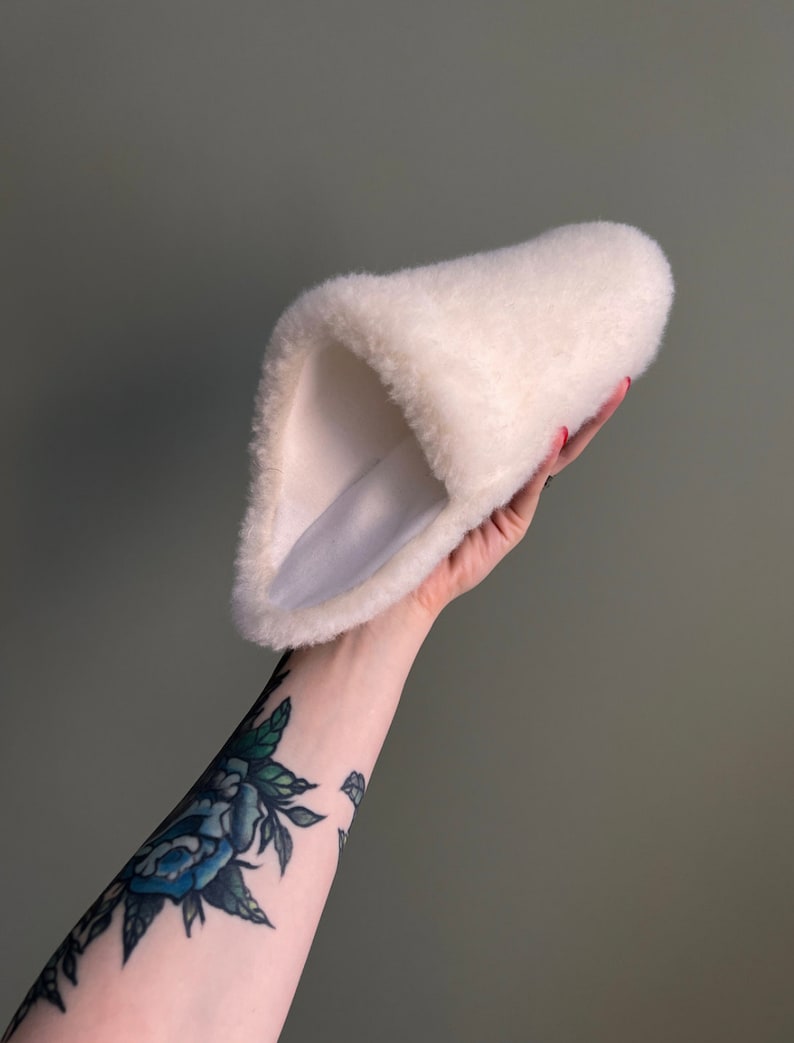 May include: A white fluffy slipper with a soft, plush lining. The slipper is folded in half and held in a hand.