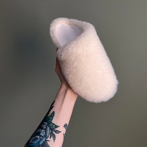 May include: A white fluffy slipper with a white lining. The slipper is being held up by a hand with a blue rose tattoo.