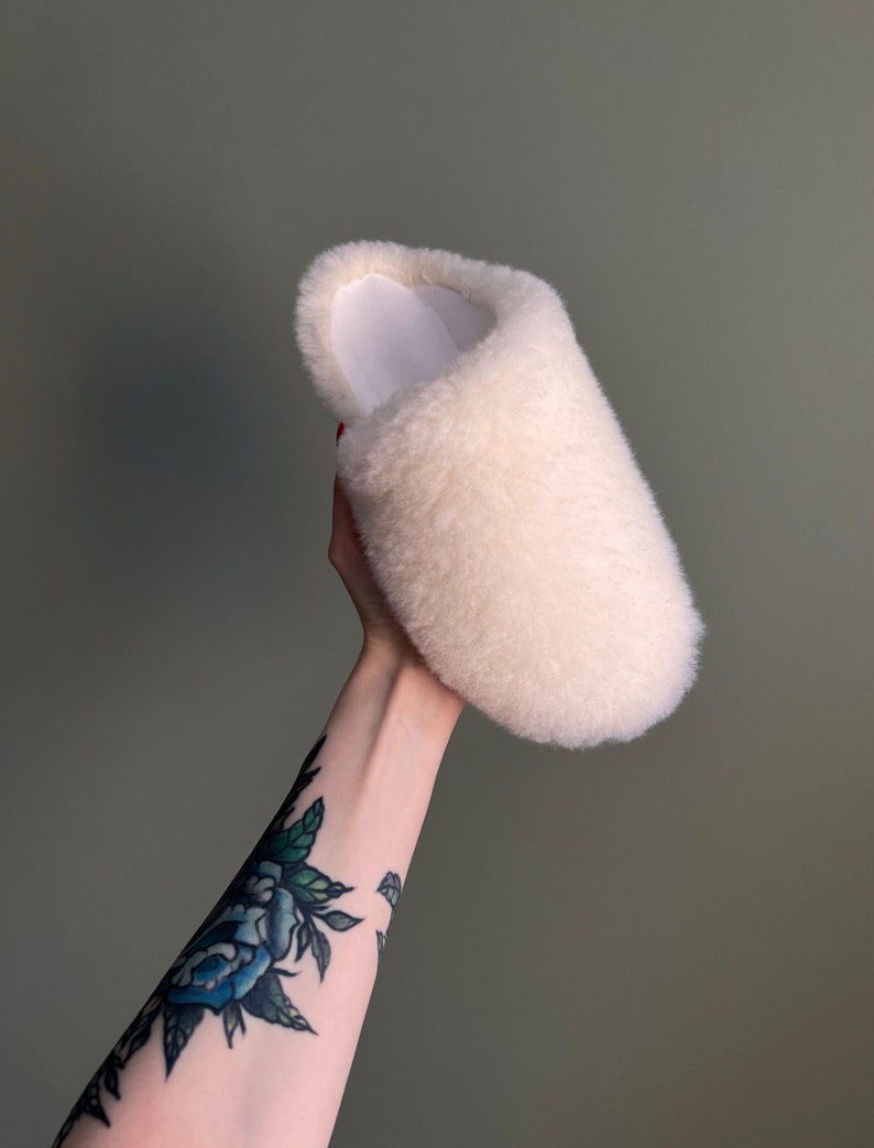 May include: A white fluffy slipper with a white lining. The slipper is being held up by a hand with a blue rose tattoo.