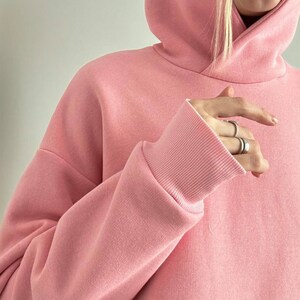 May include: A light pink hoodie with a drawstring hood and ribbed cuffs. The sweatshirt is made of a soft, thick material. The sleeves are long and the fit is oversized.