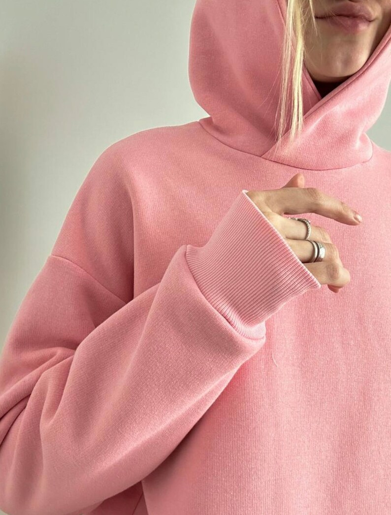 May include: A light pink hoodie with a drawstring hood and ribbed cuffs. The sweatshirt is made of a soft, thick material. The sleeves are long and the fit is oversized.