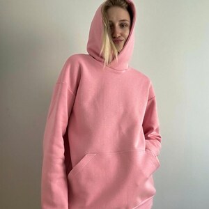 May include: A light pink hoodie with a kangaroo pocket. The hooded sweatshirt is made of a soft material and has long sleeves. The person in the image is wearing the hoodie. The background is a plain white wall.