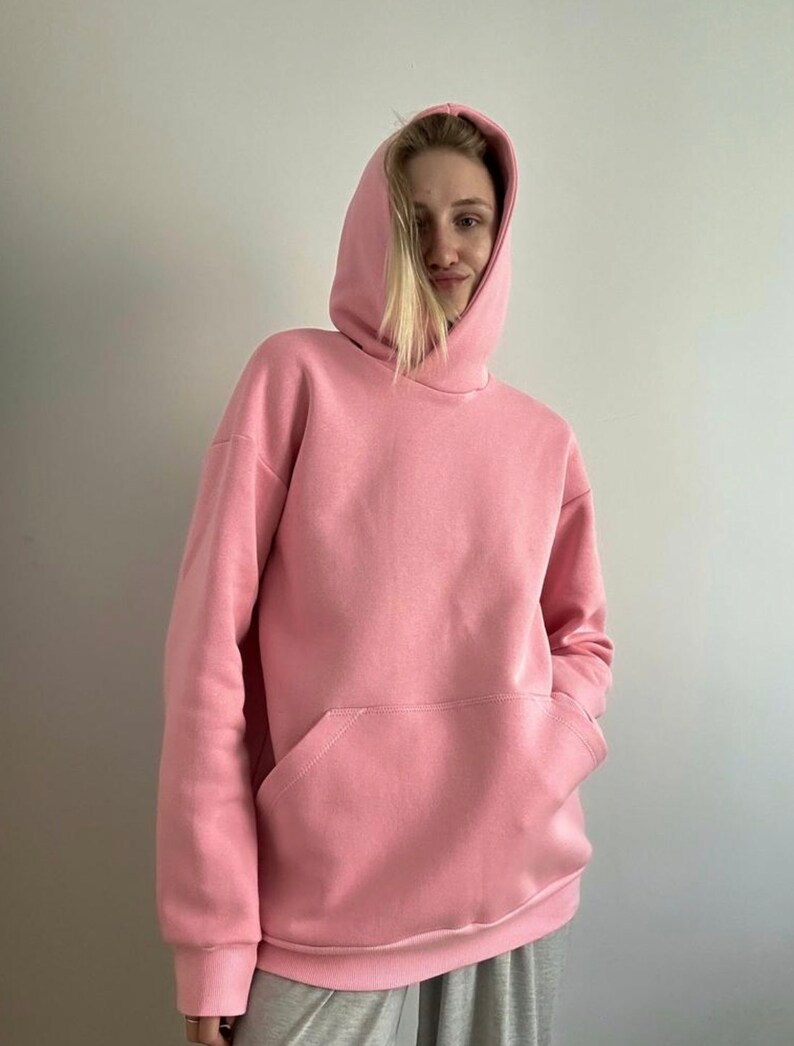 May include: A light pink hoodie with a kangaroo pocket. The hooded sweatshirt is made of a soft material and has long sleeves. The person in the image is wearing the hoodie. The background is a plain white wall.