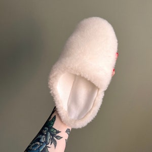 May include: White fluffy sheepskin slipper with a soft, white lining. The slipper is held in a hand with a tattoo.