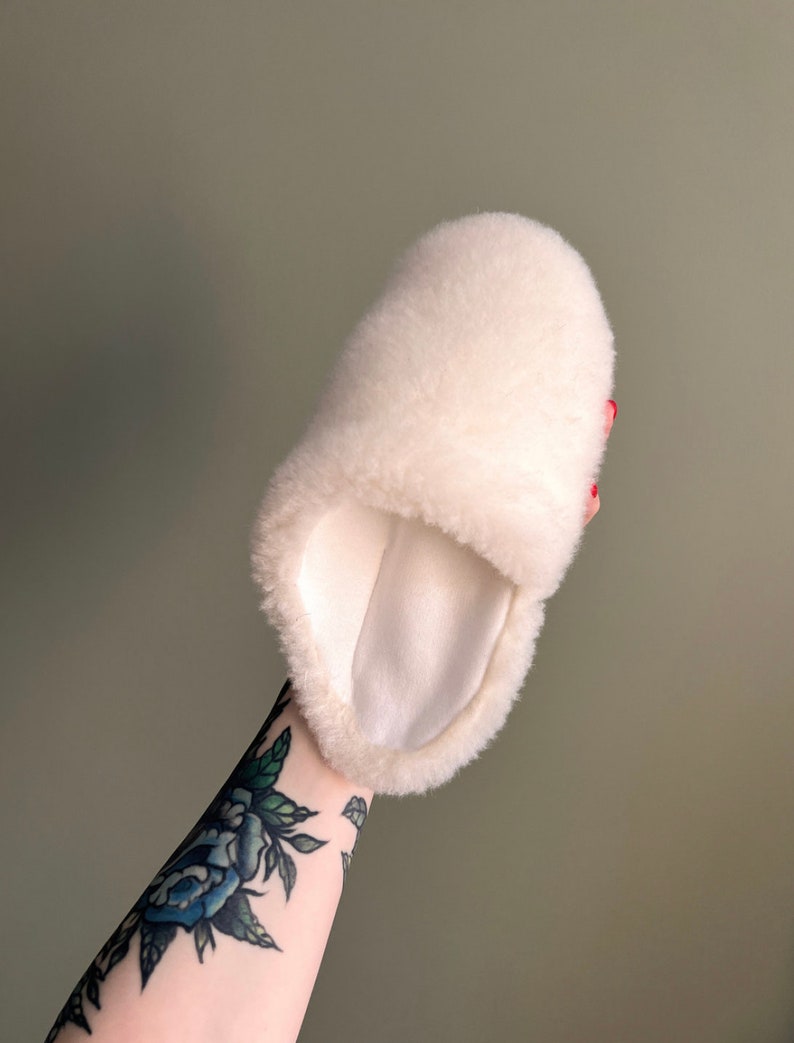 May include: White fluffy sheepskin slipper with a soft, white lining. The slipper is held in a hand with a tattoo.