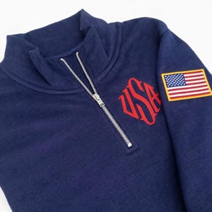 May include: Navy blue sweatshirt with a quarter-zip and a stand-up collar. It has a red "USA" monogram on the chest and an American flag patch on the upper sleeve. The sweatshirt is made of a soft, comfortable material.