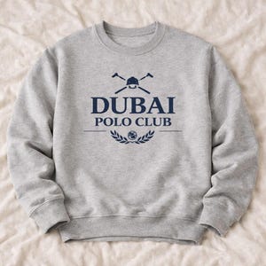 Pode incluir: Um moletom cinza claro com o texto "DUBAI POLO CLUB" em azul marinho. O design inclui um capacete de polo e tacos cruzados acima do texto e uma coroa de louros abaixo. O moletom tem gola redonda e mangas compridas.