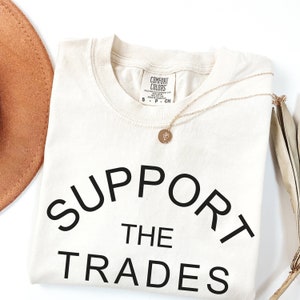 May include: A cream colored t-shirt with the text "SUPPORT THE TRADES" in black lettering. The shirt is folded and laid flat on a white surface.