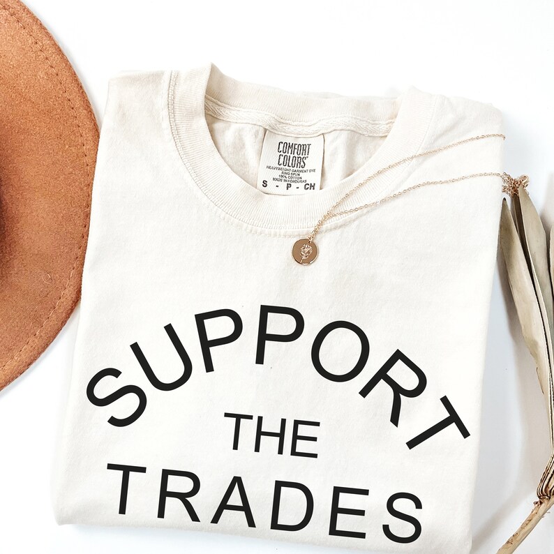 May include: A cream colored t-shirt with the text "SUPPORT THE TRADES" in black lettering. The shirt is folded and laid flat on a white surface.