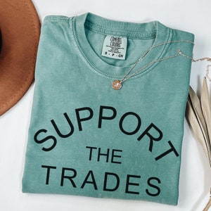 May include: A teal blue t-shirt with the text "SUPPORT THE TRADES" in black lettering. The shirt is made of a soft, comfortable material.